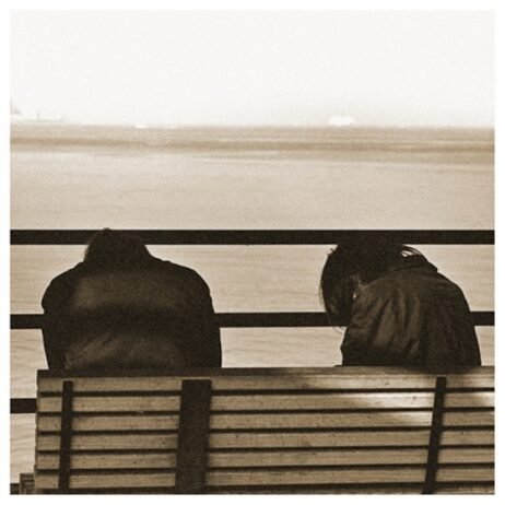 The image is the album cover for Metz - II. The cover features a sepia-toned photograph of two figures sitting on a bench, facing away from the viewer, looking out over a body of water. Their heads are slumped down. The image is minimalistic and muted, with the figures' backs emphasized against the simple, peaceful backdrop of the sea and horizon.