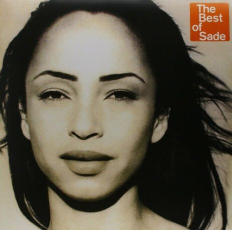 The image is the album cover for "The Best of Sade." It features a close-up, sepia-toned portrait of Sade Adu, looking directly into the camera with a calm expression. Her hair flows slightly in the wind, adding a sense of movement to the serene image. In the upper right corner, a red square contains the text "The Best of Sade" in white letters.