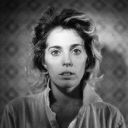 The image is a black-and-white portrait of a woman with a neutral expression. Her hair is slightly tousled, and she wears a collared shirt, partially unbuttoned. The background is softly blurred with a subtle pattern, which helps draw focus to her face. The lighting and shadows give the photograph a classic and raw aesthetic, evoking a sense of introspection and vulnerability.