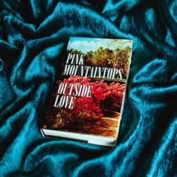 The image features a book or album cover placed on a luxurious, textured blue fabric background. The cover displays a photograph of a scenic outdoor setting with bright pink flowering bushes and lush greenery.
