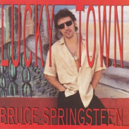 This image is the album cover of "Lucky Town" by Bruce Springsteen. It shows Bruce Springsteen leaning casually against a wall with his hands in his pockets, wearing sunglasses, a light beige shirt, and a necklace. The title "Lucky Town" is written in large, bold red letters at the top, while "Bruce Springsteen" appears at the bottom in the same bold red font.