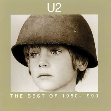 The album cover for U2's - The Best of 1980-1990 features a sepia-toned photograph of a young boy wearing a helmet, looking contemplative and innocent. The band's name "U2" is displayed at the top in a simple, white font, while the album title is positioned at the bottom in all capital letters. The image is set against a muted beige background.