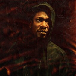 The image appears to be an album cover featuring Roots Manuva. The artwork has a dark, moody atmosphere, showing Roots Manuva wearing a hooded jacket. His expression is serious, and the lighting casts dramatic shadows across his face, adding to the intensity of the image. The background has a textured, reddish-brown hue, which gives a fiery and layered effect, enhancing the overall feeling of depth and mystery.