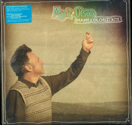 The image is the album cover for Mark Olson - Many Colored Kite. The cover shows Mark Olson standing outdoors in a pastoral setting, gazing upwards with his arm raised as if he's about to release something into the sky. The design has a soft, vintage aesthetic, with muted colours and a subtle texture overlay. The album title "Many Colored Kite" is written in a whimsical, playful font next to the artist's name "Mark Olson."