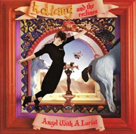 This image is the album cover for "Angel with a Lariat" by k.d. lang and the Reclines. The design features a colorful scene, with k.d. lang posed in the center, surrounded by various eclectic elements.. At the top, a pink banner reads "k.d. lang and the reclines" in a yellow-and-red font, while the album title, "Angel With A Lariat," appears at the bottom in red text on another ribbon-like banner.