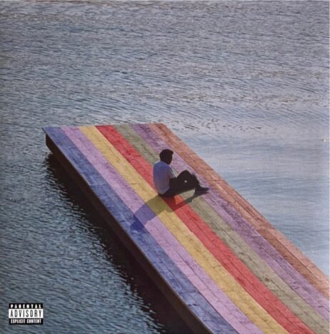 The album cover depicts a person sitting alone on a floating wooden dock, painted in vibrant, rainbow-colored stripes. The dock is positioned in the middle of a calm, expansive body of water, creating a sense of isolation and introspection. The figure is facing away from the viewer, looking out over the water, adding to the contemplative mood. The image is minimalistic, with the vast water and the solitary figure contrasting against the bright, colorful dock. The lower-left corner features a small "Parental Advisory" label.