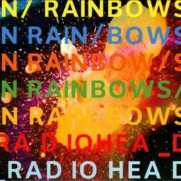 The cover displays the title "In Rainbows" repeatedly in different colors: yellow, blue, red, and green. The text is set against a vibrant, abstract background with splashes of colors, including orange, yellow, pink, and black, giving it a cosmic or explosive appearance. The band name "Radiohead" is displayed at the bottom in a similar multi-colored format.