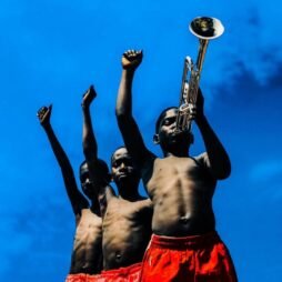 This is the album cover for Common - Revolution. It features three young boys under a bright blue sky, each raising a fist in the air. The boy in the foreground holds a trumpet to his lips, symbolizing music and resilience. They are all shirtless, wearing red shorts.