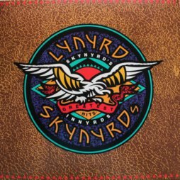 The album cover for Lynyrd Skynyrd's Greatest Hits features a stylized circular emblem with a large eagle spreading its wings in the center. The eagle is set against a vibrant background with red, blue, and green hues. The band's name, "Lynyrd Skynyrd," is prominently displayed in a bold, colorful font encircling the eagle, with "Greatest Hits" written across the bottom. The background has a rustic, textured pattern.