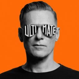 A monochrome image of Bryan Adamâ€™s - Ultimate album cover consists of a man wearing reflective sunglasses with the word "ULTIMATE" written across the lenses in bold, block letters. The background is a vibrant, solid orange, creating a stark contrast. The man has a serious expression, with neatly styled hair, and is dressed in a plain black shirt, standing against the bright background.