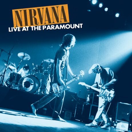 A black-and-white concert photo of Nirvana performing on stage. The image is tinted in blue with Kurt Cobain in the center, holding his guitar, and Krist Novoselic on the right, leaning towards his bass guitar. The drummer is visible in the background behind a drum set. The band's name "NIRVANA" appears in bold yellow letters in the top left corner, and the album title "LIVE AT THE PARAMOUNT" is in white text below it.