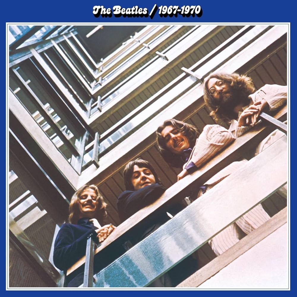 The album cover for The Beatles 1967–1970 features an iconic photograph of The Beatles taken from a low angle. The members—John Lennon, Paul McCartney, George Harrison, and Ringo Starr—are looking down from a balcony. The shot creates a symmetrical, geometric effect with the building's structure. The album title is presented at the top in a simple white font against a blue background.