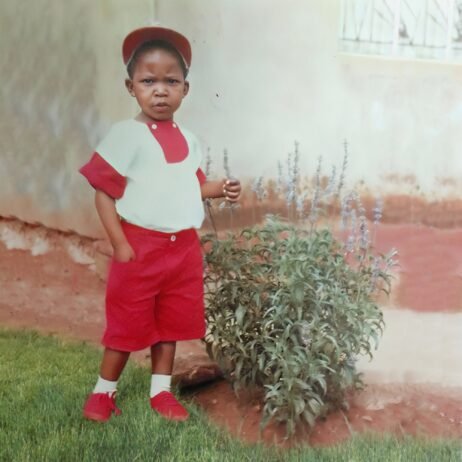 This is the album cover of Malcolm Jiyane Tree-O - True Story. A young child stands in a yard, dressed in a red and white outfit with matching red shorts, shirt, hat, and shoes. The child has a serious expression and stands beside a small green plant with purple flowers. The background shows a wall with a faded white color and a patch of red soil.