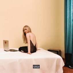 This is the album cover for Sabrina Carpenter - Emails I canâ€™t send. A woman with long blonde hair sits on a bed in a dimly lit room. She is wearing a black, backless dress and looking over her shoulder at the camera. Beside her is a laptop with a red heart sticker. The room features minimalist decor with plain beige walls and teal curtains.