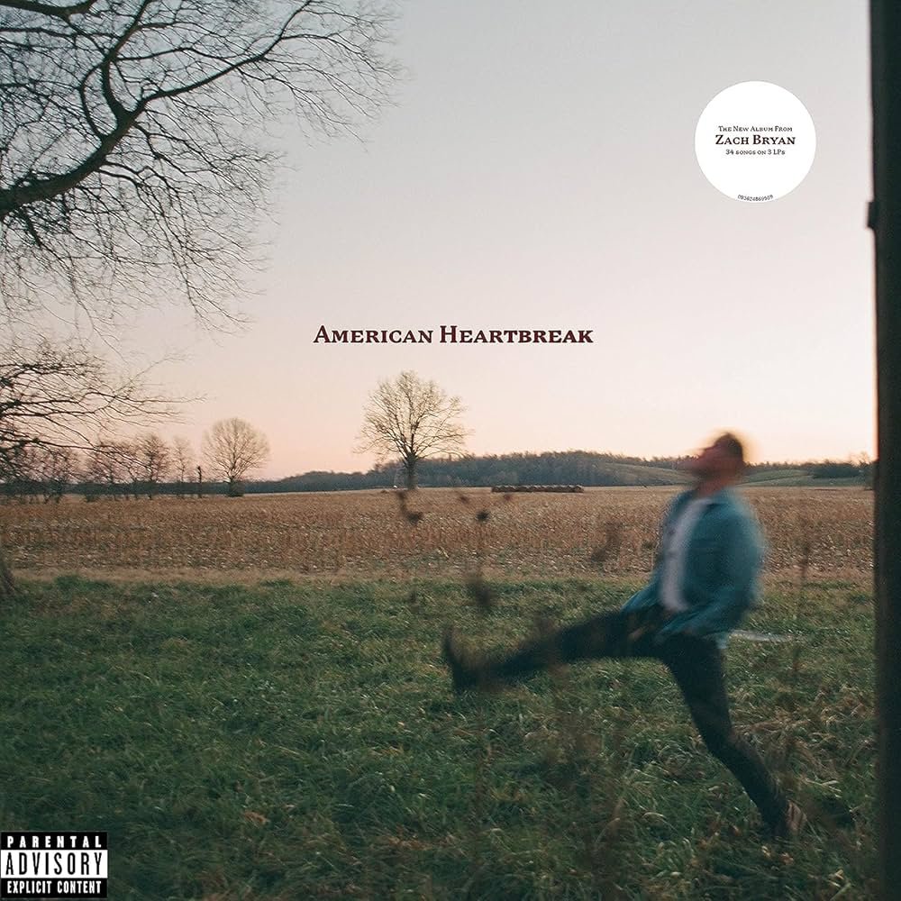 The album cover features a blurred image of a man kicking up leaves in a vast, empty field at dusk. Bare trees and the soft pink and orange sky create a serene, melancholic atmosphere. The title "American Heartbreak" is centered in simple black text, with Zach Bryan's name in a white circle above.