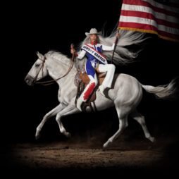This is the album cover for BeyoncÃ© - Cowboy Carter. It features BeyoncÃ© dressed in a red, white, and blue cowgirl outfit and rides a white horse in motion. She holds a large American flag, and her long, flowing silver hair trails dramatically behind her.