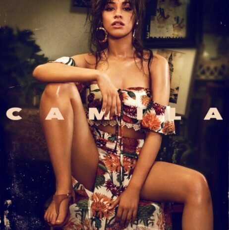 The image is the album cover for "Camila" by Camila Cabello. The cover features a photograph of Camila Cabello sitting in a relaxed pose. She is wearing a floral two-piece outfit with an off-the-shoulder top and a high-slit skirt. Her hair is slightly tousled, and she wears large hoop earrings. The album's title, "CAMILA," is displayed in bold, white capital letters across the middle of the image, directly over her legs.