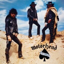 This is the album cover for MotÃ¶rhead - Ace Of Spades. It features 3 men in black leather cowboy attire standing on a sandy mountain surface. The background is of a blue cloudy sky.